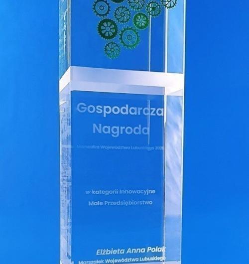szklana statuetka zębatki nagroda gospodarcza Marszałka Województwa Lubuskiego w kategorii innowacyjne przedsiębiorstwo. Statuetka kryształowy blok, połączenie trzech technik zdobienia szkła, grawerowanie 3D, piaskowanie ozdobnych pasków oraz malowanie. Winogrona symbol Zielonej Góry i zębatki symbol innowacyjności są w pięciu kolorach pasujących do kategorii w konkursie