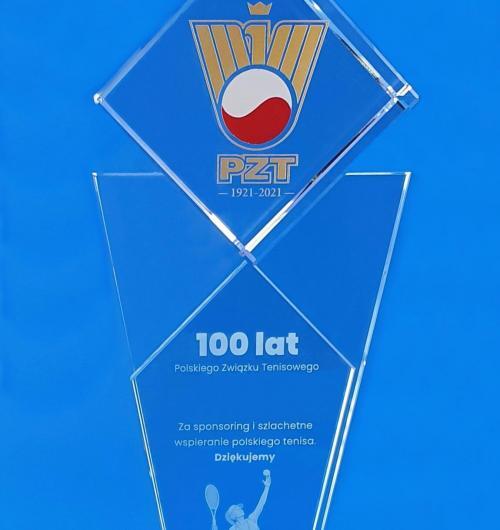 prestiżowa szklana statuetka, puchar sportowy z okazji 100-lecia PZT Szklana statuetka z jubileuszowym logo w kryształowym sześcianie Szklane podziękowanie za wspieranie sportu z okazji 100-lecia Polskiego Związku Tenisowego Oprócz dedykacji na dole statuetki jest wygrawerowana laserem 3D tenisistka lub tenisista z rakietą w trakcie uderzenia w piłkę Okazała szklana statuetka na 100 lat polskiego tenisa