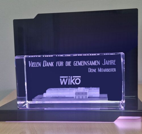 szklana statuetka z budynkiem firmy w 3D umieszczona na eleganckiej czarnej, podświetlanej podstawce z diodami LED Budynek wygrawerowany w kryształowej bryle szkła jako obiekt trójwymiarowy Szklana statuetka z budynkiem 3D to prezent dla właściciela firmy z okazji przejścia na emeryturę Ekskluzywny szklany prezent jest w zestawie z podświetlaną podstawką na pilota, dzięki któremu można dowolnie zmieniać kolory podświetlenia Jeżeli szukasz eleganckiego i nowoczesnego, personalizowanego prezentu firmowego zapytaj u nas, już od 25 lat zajmujemy się grawerowaniem 3D Laser 3D producent szklanych statuetek z Zielonej Góry