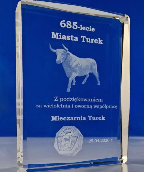 szklana statuetka z Turem w 3D z okazji 685-lecia miasta Turek od Mleczarni Turek Elegancka szklana statuetka z ozdobnymi szlifami po bokach, które pięknie załamują światło i dodają statuetce blasku. Statuetka ze szkła kryształowego z grawerem 3D z okazji 685-lecia lokacji miasta Turek jest prezentem dla władz miasta. To bardzo elegancka nagroda okolicznościowa z okazji rocznicy miasta W statuetce jest wygrawerowany trójwymiarowy Tur – symbol miasta. Szklana statuetka z turem i jubileuszową dedykacją to upominek od Mleczarni Turek w podziękowaniu za wieloletnią i owocną współpracę. Oprócz tura w statuetce jest wygrawerowane okolicznościowe opakowanie sera Camembert. Statuetka wykonana przez producenta szklanych statuetek z Zielonej Góry, firmę Laser 3D Laser 3D to pionier grawerowania 3D w Polsce