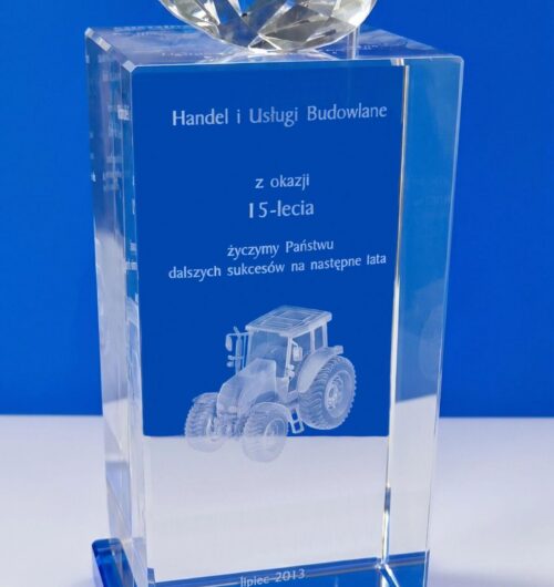 spersonalizowana-szklana-statuetka-z-grawerem-3d-traktor-15-lecie spersonalizowana szklana statuetka z grawerem 3D traktora z okazji 15-lecia firmy Kryształowa statuetka z wysokiej jakości szkła kryształowego z dekoracyjnym elementem na górze w kształcie diamentu oraz z niebieską, szklaną podstawą Wewnątrz szkła metodą laserową jest wygrawerowany trójwymiarowy model ciągnika, co czyni ją idealnym upominkiem dla rolników lub firm związanych z rolnictwem Statuetka została wykonana przez producenta statuetek firmę Laser 3D z Zielonej Góry Laser 3D to pionier grawerowania 3D w szkle w Polsce. Grawerujemy już ponad 25 lat