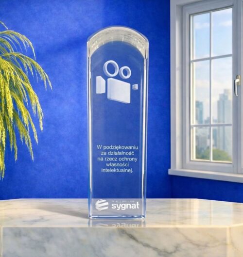 szklana-statuetka-kamera-3d-nagroda-biznesowa-grawer Statuetka w formie masywnego, kryształowego obelisku o wadze 1,6 kg z zaokrągloną górą. Wewnątrz szkła znajduje się grawer 3D przedstawiający kamerę filmową oraz napis z podziękowaniem za działalność na rzecz ochrony własności intelektualnej. Na dole widnieje logo Sygnał oraz napis 25 lat i daty.